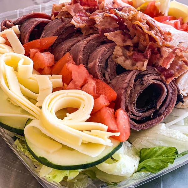 A platter of rolled deli meats, crispy bacon, diced tomatoes, cheese ribbons, and sliced cucumbers.