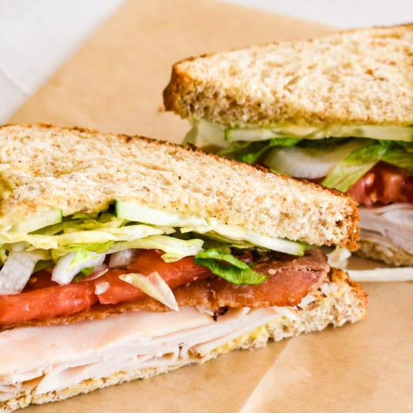 Two All American Heroes sandwiches with turkey, bacon, lettuce, tomato, and onion on whole wheat bread.