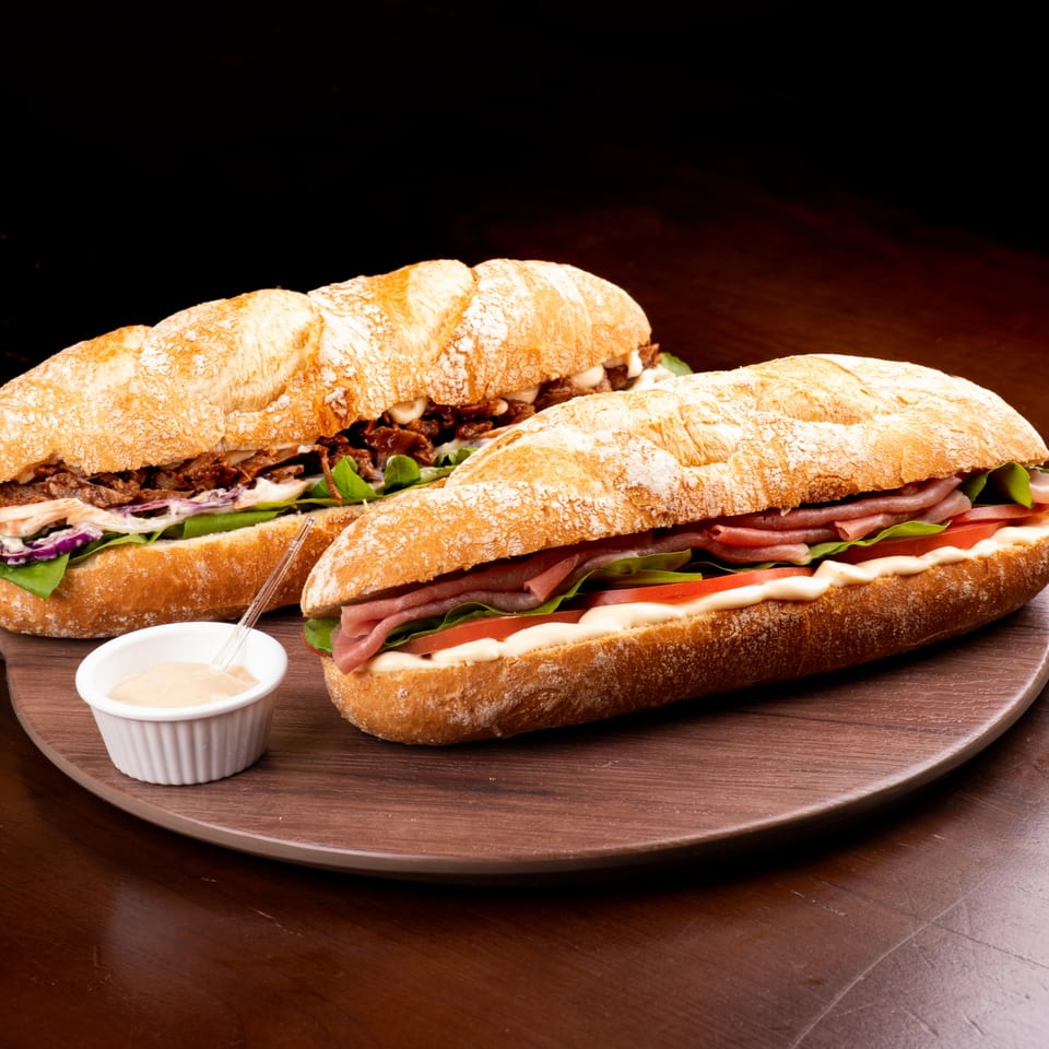 Two All American Heroes sandwiches on a wooden board, one with ham and the other with shredded meat.
