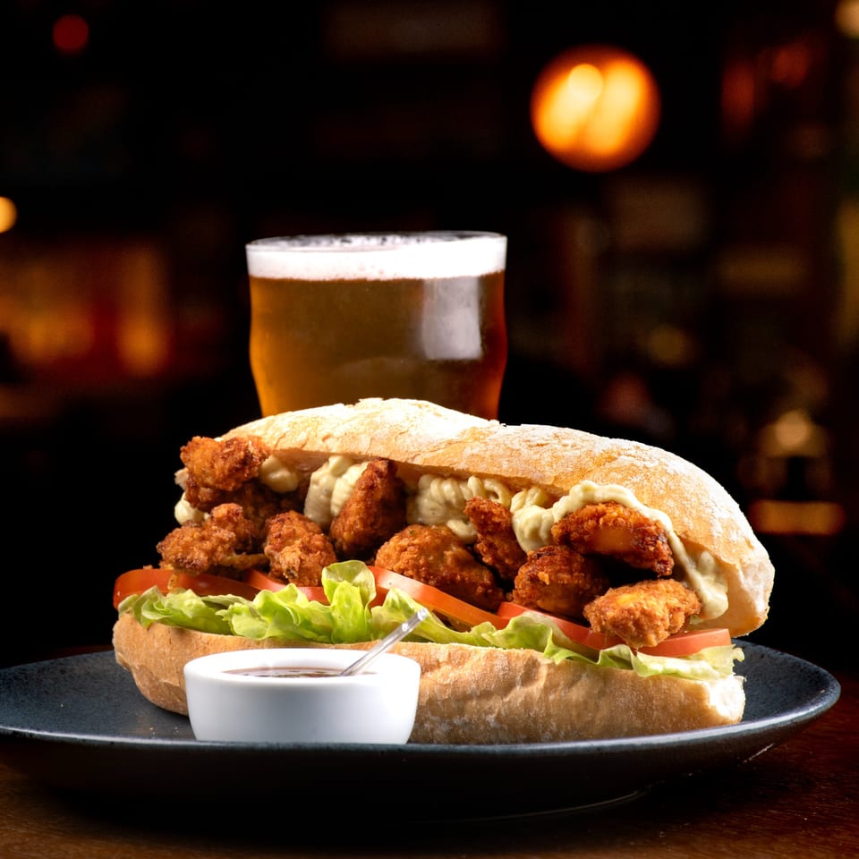 Crispy fried shrimp po'boy sandwich with lettuce, tomato, and sauce, served with a beer.