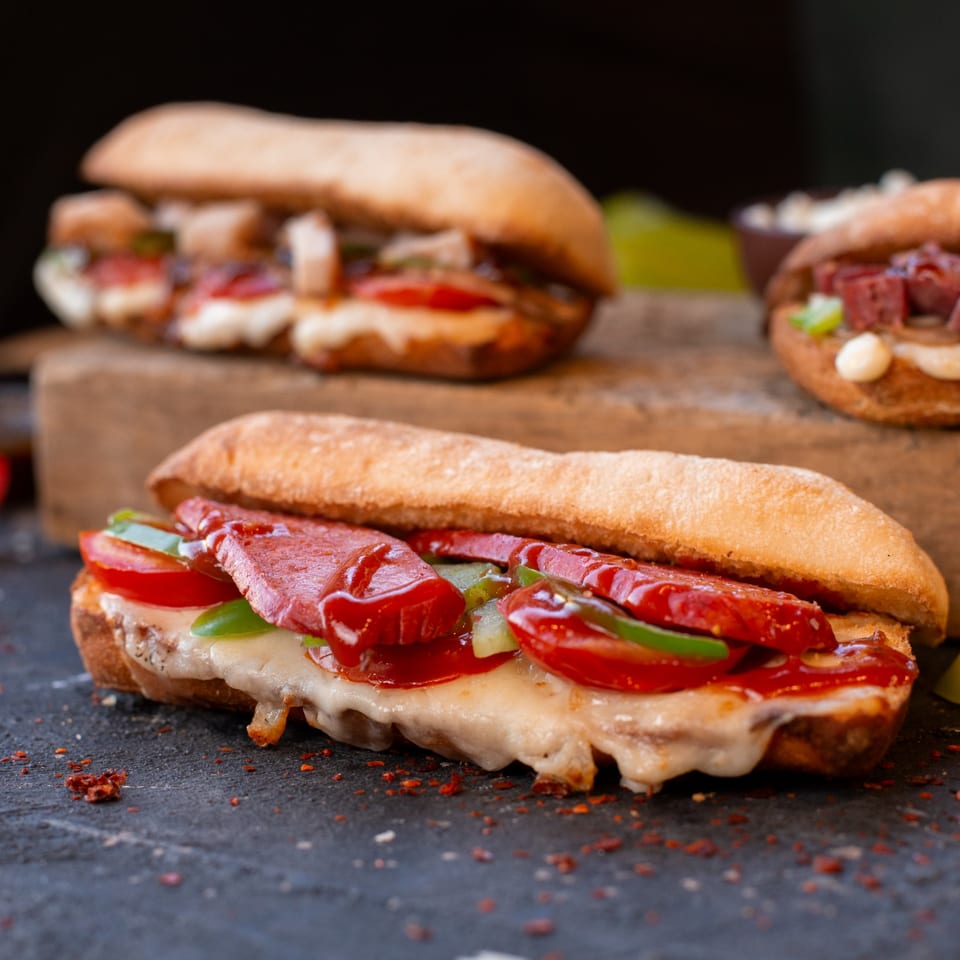 Three All American Heroes sandwiches, one in focus with melted cheese, pepperoni, tomatoes, and green peppers.