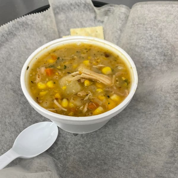A white bowl of chicken and corn chowder with a spoon and cracker on a paper-lined tray.